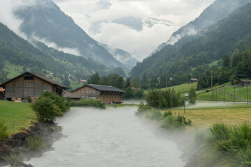 View of an alpine river in the valley during a rainy summer day. Mountains on all sides, clouds hanging between the mountains. Spray is coming of the wild river. Mystic landscape in Switzerland.