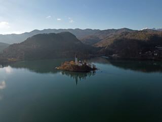 Obraz premium Lake Bled island with sun rays in the snowless winter aerial view. Ojstrica mountain and Julian Alps in the background. Perfect reflection of the island and the church on calm water surface.