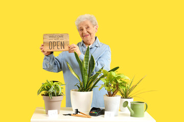 Senior florist with OPEN sign and plants on counter against yellow background
