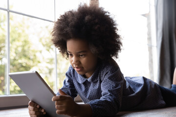 Cute mixed-race African girl lying on floor indoor with digital tablet. Little 6s kid holds modern...