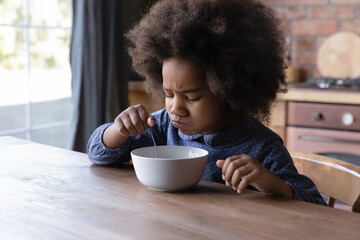 Little 6s cute African curly-haired girl sits at dining table in kitchen frowning while eats not...