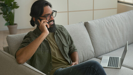 Obraz premium Muslim Arabian guy freelancer sit on couch with laptop talk mobile phone at home smiling Indian man businessman in eyeglasses speak smartphone answer call carefree converse cell business discussion
