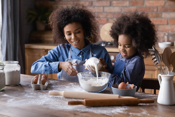 African woman teach little cute daughter to cook, family enjoy cookery, pour milk into bowl preparing for breakfast pancakes for family. Hobby, development of child, share skills, motherhood concept
