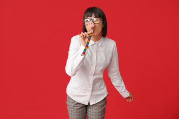 Funny young businesswoman with party blower on red background. Fool's day