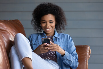 Relaxed African 30s woman sit on sofa with smartphone. E-commerce retail services user buy online...