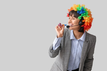 Funny young female technical support worker with headset and party blower on grey background. Fool's day