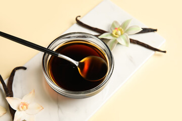 Vanilla extract in bowl with spoon, vanilla pods and flowers on light beige background