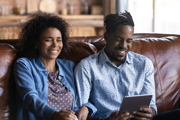 Laughing African couple sit on sofa with digital tablet, feels overjoyed have fun watch comical...