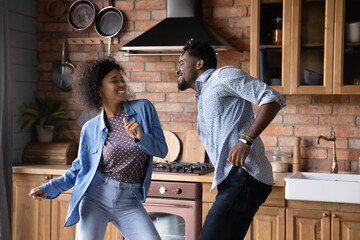 Lively millennial African hipster couple dancing in kitchen at home. Vivacious wife and husband enjoy weekend, listen favourite music moving indoor looking overjoyed, homeowner family leisure concept