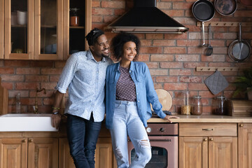 Dreamy African couple standing in modern cozy kitchen smile looking into distance. Happy homeowners family portrait, bank mortgage and tenancy, visualize bright future at new home, daydreams concept