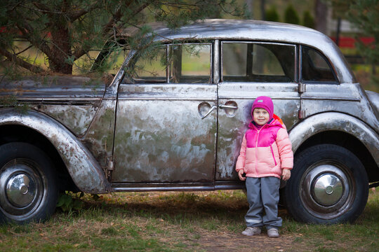 A little girl is standing by a vintage silver car outdoors in fall. Pink and gray autumn kid's outfit. Horizontal image.
