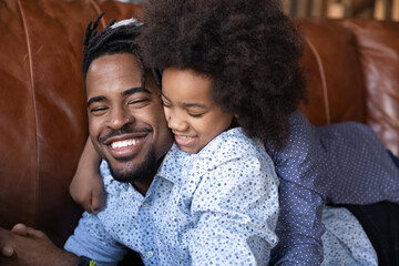 Close up smiling faces African dad and cute daughter enjoy playtime, carefree time together lying on sofa spend weekend at home, playing feeling love and bond. Loving family, happy fatherhood concept