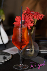 A glass with a cool Aperol cocktail stands on a table in a restaurant against a background of red flowers, a romantic date