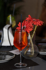 A glass with a cool Aperol cocktail stands on a table in a restaurant against a background of red flowers, a romantic date