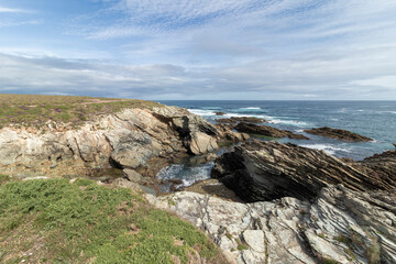 A rocky coastline with greenery, under a partly cloudy sky, waves crashing against the rocks, creating a serene atmosphere