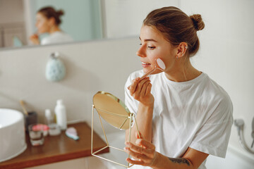 Smiling woman massaging her face with jade roller massager and looking at round cosmetic mirror