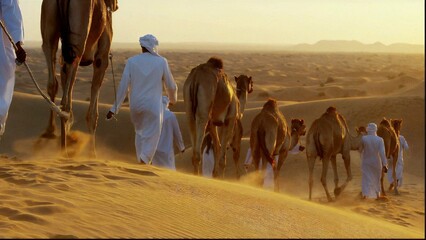 A camel caravan journeys through the desert, accompanied by Saudi people dressed in traditional white attire.