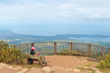 Side view of a young female sitting on bench and using smartphone at Mombacho volcano nature reserve.