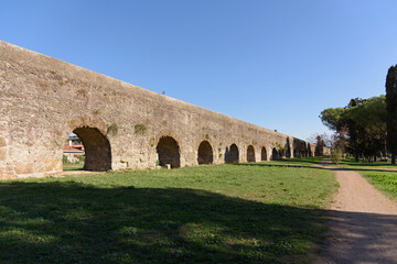 The long arched wall of an ancient Roman aqueduct along a walking path in a park, evening shadows from the setting sun shadows fall on the ground in a beautiful pattern
