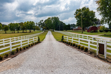 Driveway with a white picket fence and boxwoods landscaping leading to a beautiful farm.