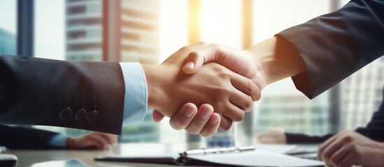 Two business professionals are engaged in a handshake gesture in an office setting. They are...