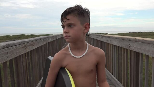 Shirtless beach boy with shell necklace walking on foot bridge carrying his boogie board