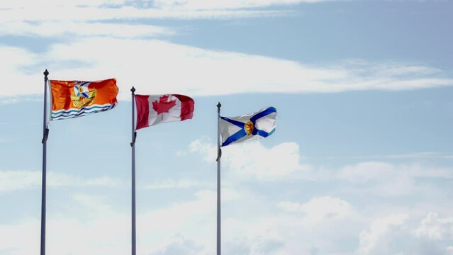 Wide shot of the Acadian flag, the Canadian flag and the Nova Scotia flag in a cascading row and blowing in the wind, against a blue sky with clouds, on a windy summer day