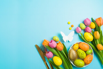 Easter holiday celebration concept with easter eggs gold cutlery and tulip flowers on blue background. Top view, flat lay composition