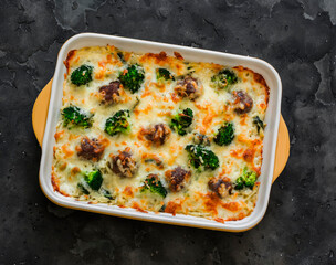 Casserole with meatballs, oregano, broccoli and cheese in a baking dish on a dark background, top view