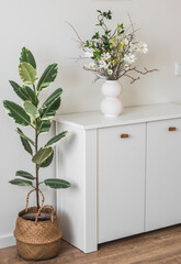 A white wooden chest of drawers with a floral arrangement in a ceramic vase, a homemade ficus in a basket in the hallway interior