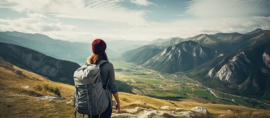 A female tourist with a backpack is standing on a mountainside, gazing at the majestic mountain peaks in the background. She appears to be taking in the awe-inspiring view. - obrazy, fototapety, plakaty