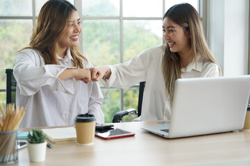 Fototapeta premium Two young Asian women roommates working at home with laptops and hand bump together with friends smiling, happy at successful online business apartment.