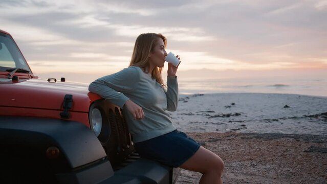 Young woman on vacation standing by car at beach watching morning sunrise and drinking coffee on road trip  - shot in real time