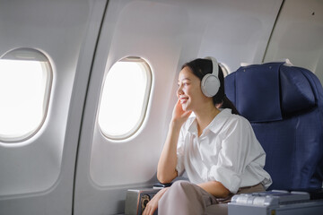 Fototapeta premium Asian woman sitting and using a smartphone in an airplane Listening to music next to the airplane seat window travel and technology concept