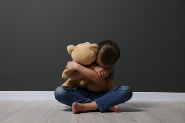 Child abuse. Upset girl with toy sitting on floor near grey wall - obrazy, fototapety, plakaty