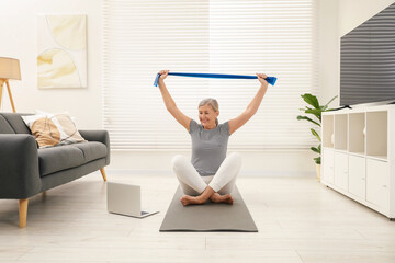 Fototapeta premium Senior woman doing exercise with fitness elastic band near laptop on mat at home