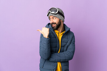 Skier man with snowboarding glasses over isolated purple background pointing to the side to present a product