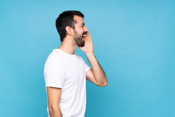 Young man with beard  over isolated blue background shouting with mouth wide open to the lateral