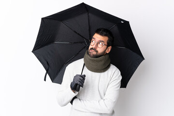 Caucasian handsome man with beard holding an umbrella over isolated white wall with confuse face...