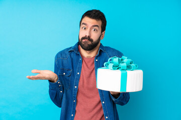 Young handsome man with a big cake over isolated blue background having doubts while raising hands