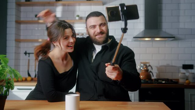 Happy young couple sitting in the kitchen taking photos with smartphone on selfie stick laughing, having fun