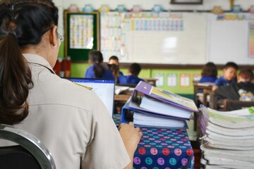 A Thai teacher is sitting and doing a lot of paperwork at a table in the classroom. While teaching at the same time.