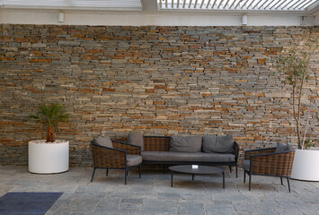 A contemporary outdoor seating area against a textured stone wall. A grey sofa and two chairs with...