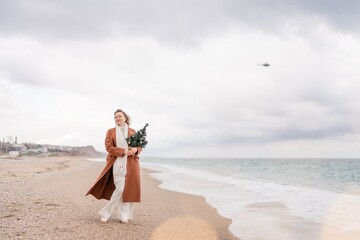 Fototapeta na wymiar Blond woman Christmas tree sea. Christmas portrait of a happy woman walking along the beach and holding a Christmas tree in her hands. She is wearing a brown coat and a white suit.
