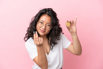 Young asian woman holding a Bitcoin isolated on pink background making money gesture