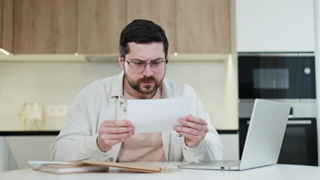 Front view of disappointed male opening packed envelope with important documents inside and feeling upset of received information. Man typing on portable laptop and trying solve problem at home.