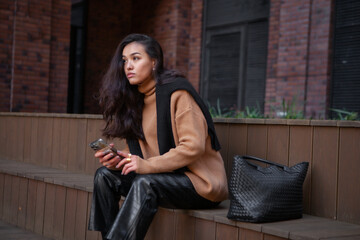 Fototapeta na wymiar Chic young brunette in fashionable layered attire beige knit sweater and leather pants sits outdoors, engrossed in her phone