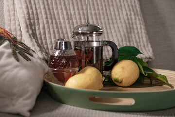 herbal tea in metal glass french press served with fresh lemon fruits , honey on wooden green tray on sofa couch with cozy decorative punch pilow, white fluffy blanket