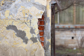 Collapsed red bricks and peeling plaster on the wall of a house with yellow walls close up in the background with a blur of part of a wooden building.