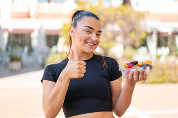 Young pretty brunette woman holding a bowl of fruit at outdoors with thumbs up because something...